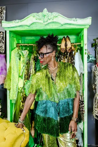 Deborah standing at the green wardrobe in a fringed jacket layered green to teal, with gold metallic trousers.